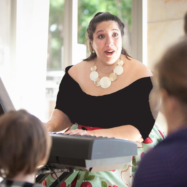 Woman wearing black dress and large gold necklace sitting at piano and singing in front of an audience. 