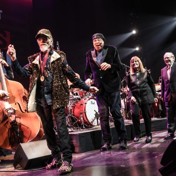 Three men and a woman smile and walk single-file onstage while musicians play behind them.