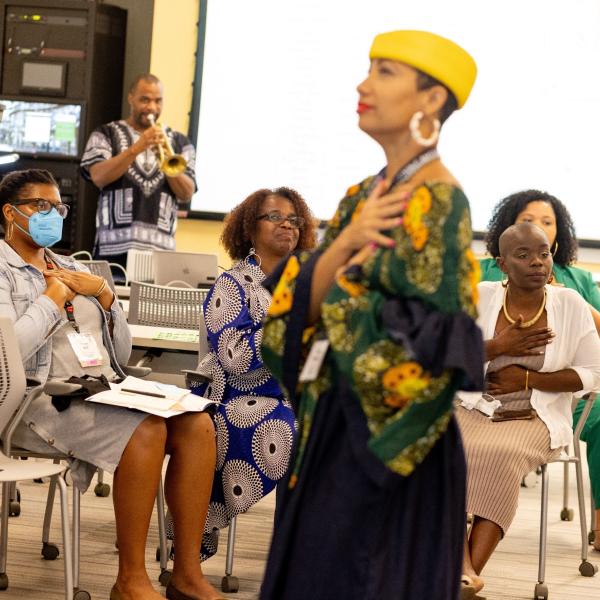 Black woman in yellow hat and flowery shirt stands in foreground with hand on her heart, group of people seated behind her also have hands over the heart, Black man at back plays a trumpet. 