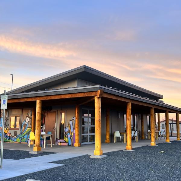 The exterior of a one story building with a colorful mural painted on one side. There are tables and chairs situated under an overhang supported by raw wood columns. The sky is filled with soft pastel pinks, blues, and yellows in what looks to be either sunrise or sunset.