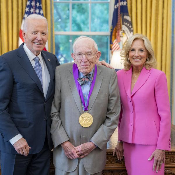 Man in gray suit wearing a medal around his neck surrounded by man wearing a blue suit and woman with blonde hair wearing a pink suit. 