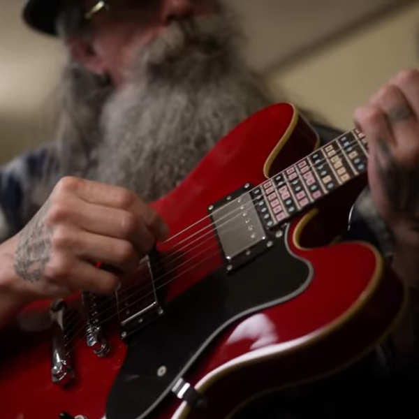 A man with a long grey beard and mustache sits behind a red guitar. His tattooed hands in the midst of playing a tune