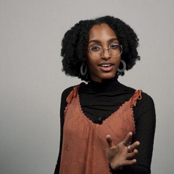 Woman in black turtleneck under orange dress gestures forward with hand while reciting to camera