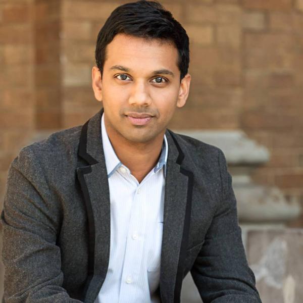 Portrait of Asian man wearing a white shirt and gray sports jacket in front of brick wall.
