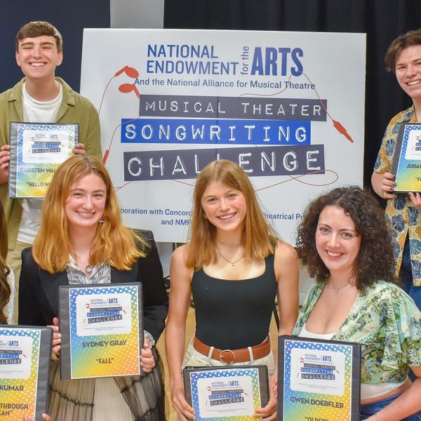 A group of teenagers smile and pose in front of a sign for the Musical Theater Songwriting Challenge. They each hold a binder with their name and the name of their winning song.