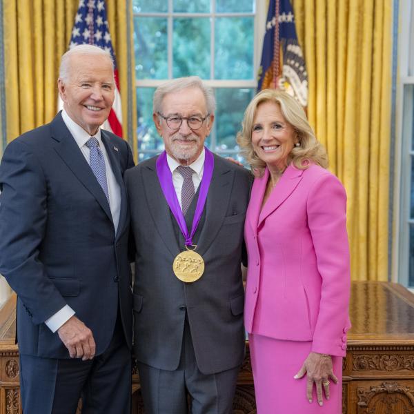 Man with glasses and gray hair and beard in gray suit wearing a medal around his neck surrounded by older man wearing a blue suit and woman with blonde hair wearing a pink suit. 