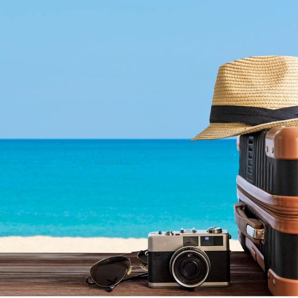 A plane flies over a beach. A cream hat is sitting on two suitcases