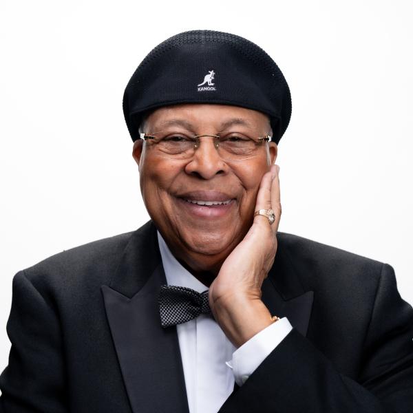 Latino man wearing tuxedo and bow-tie, with beret, left hand holding his face, smiling.