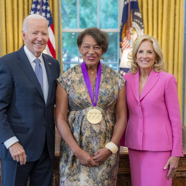 Black woman in flowery dress wearing a medal around her neck surrounded by older man wearing a blue suit and woman with blonde hair wearing a pink suit. 