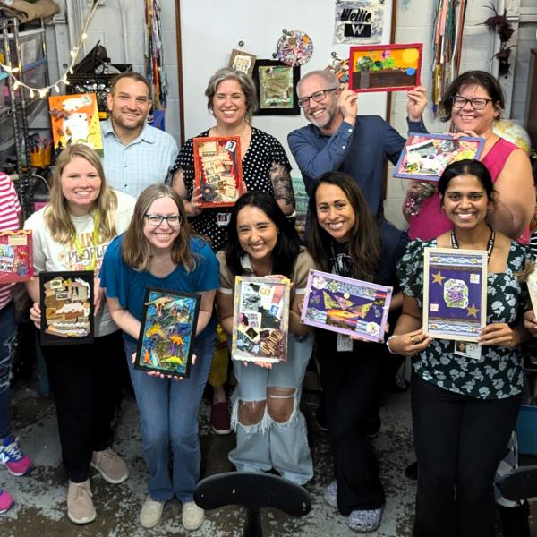 A group of teachers stand for a group photo, smiling and holding up their artwork.
