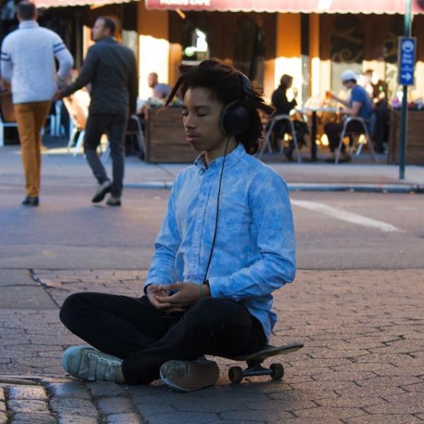 A ypoung in a meditation pose aittiing on a skate board while a number of people go about their business around him.