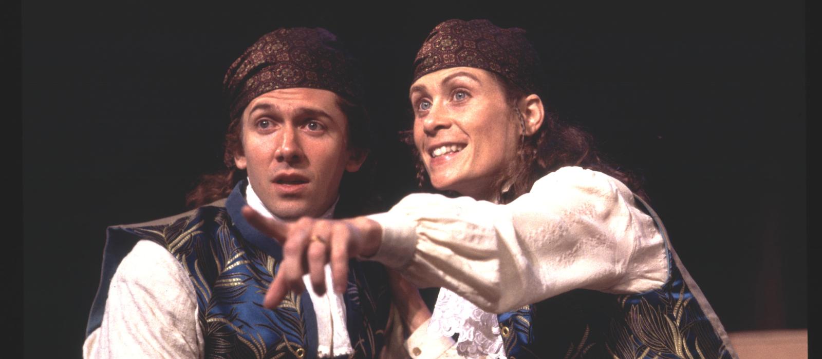 A man and a woman on stage dressed identically in white shirt, colorful vest, and cap.