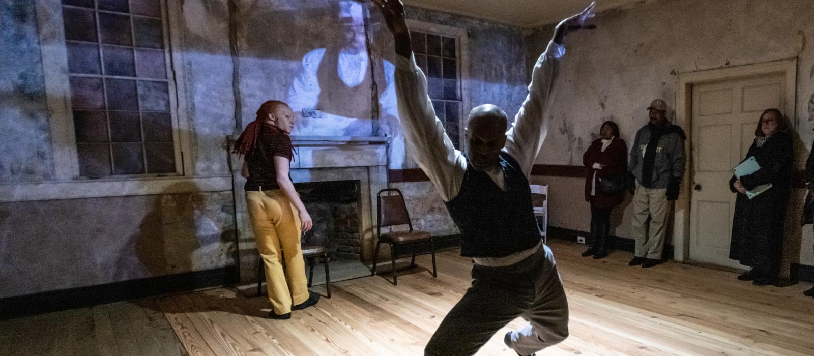 Black man in foreground dancing in room with man at wall behind him in front of projection on wall.