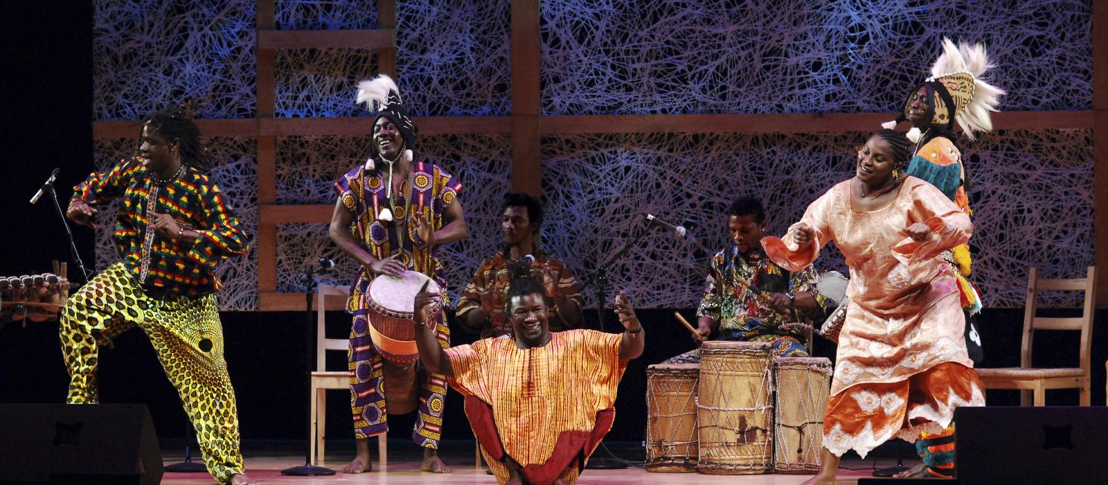 Black man in African garb with no legs dancing on stage surrounded by women in African garb also dancing