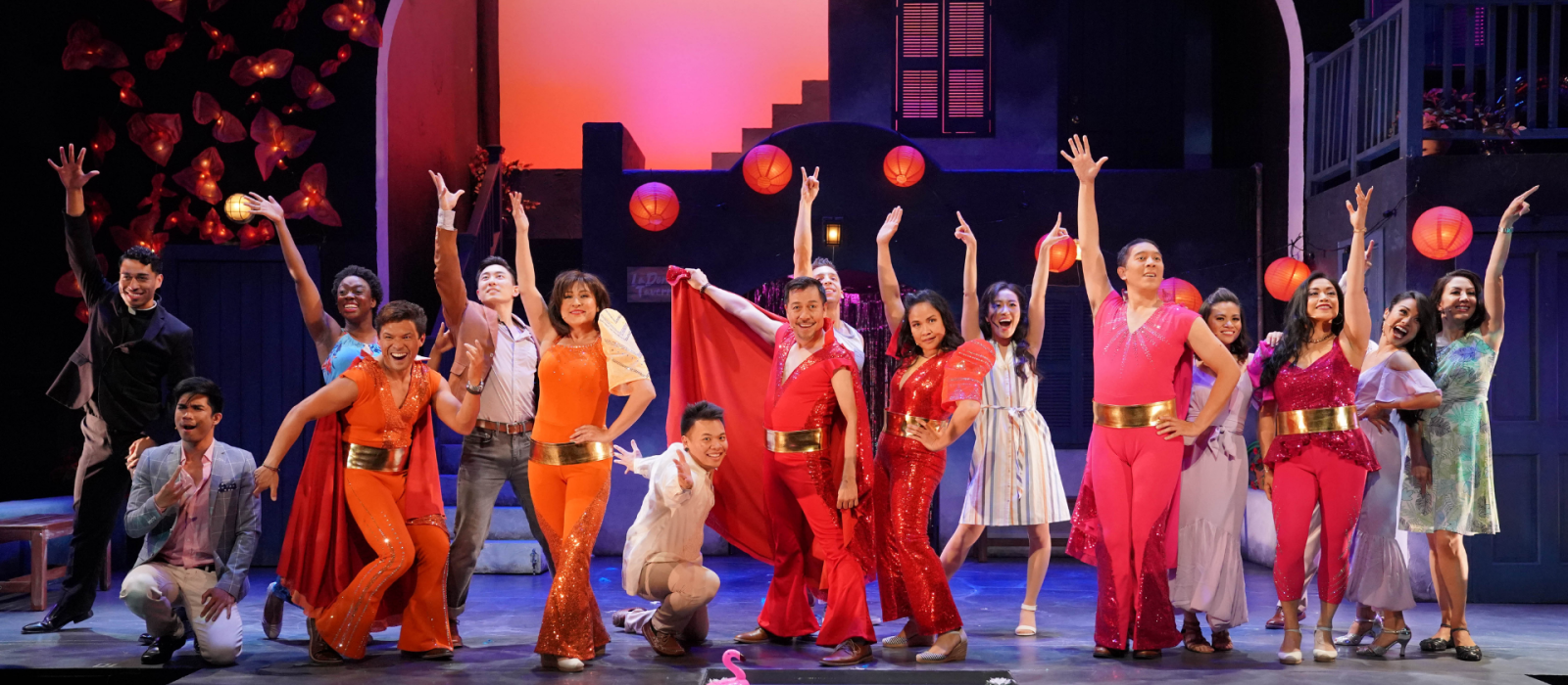 A group of Asian performers on stage, dressed in flashy mostly red costumes holding their arms in the air for a finale.