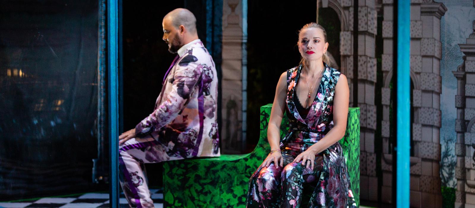 Man and woman wearing colorful clothing sitting on stage with black and white floor.