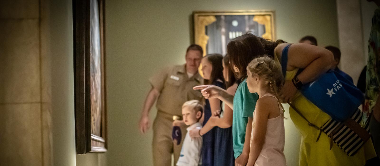 A woman in a yellow dress surrounded by children points to a painting on the wall of a museum.