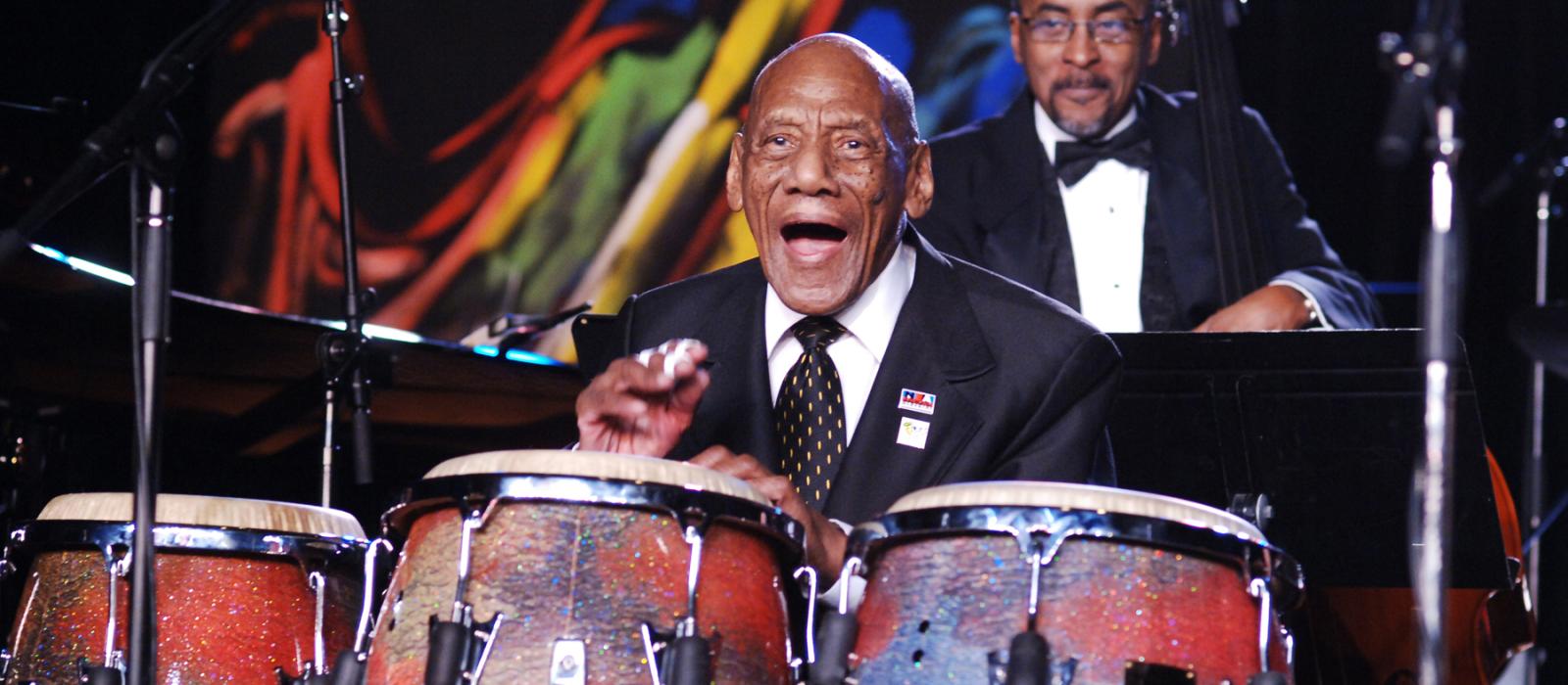 Older Hispanic man in suit playing congas on stage.