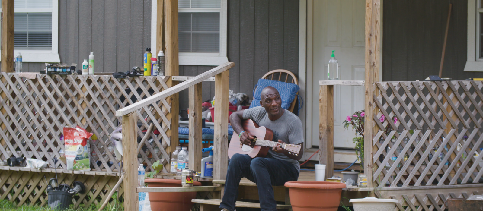 A bald Black man sitting on a porch playing a guitar