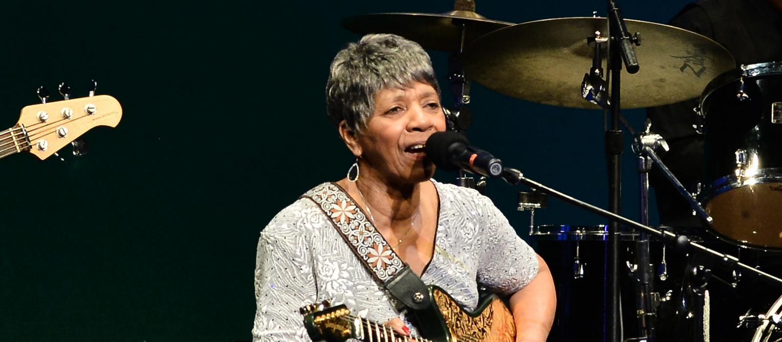 Black woman sitting on stage playing guitar left-handed.