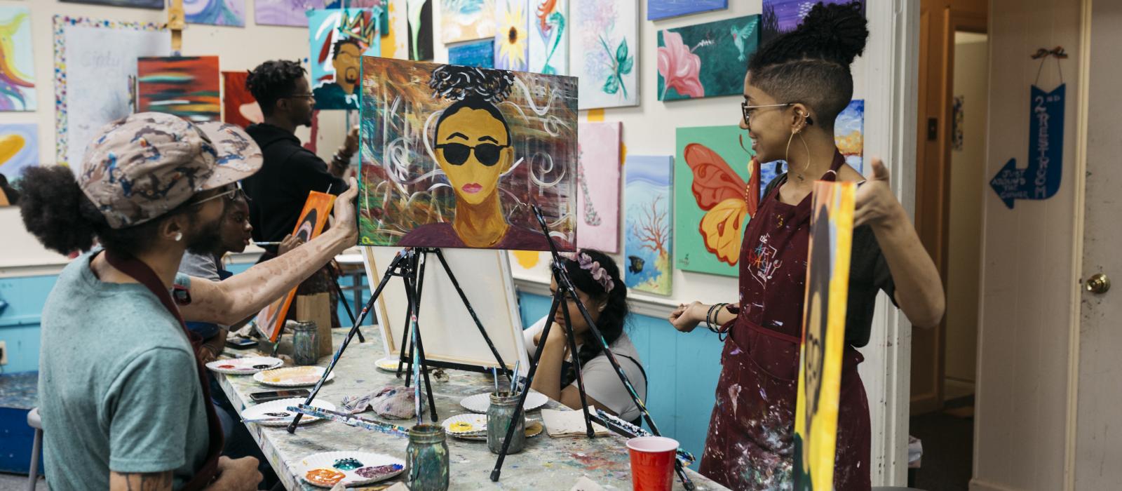 Three students in a school art room , two of the students looking at a portrait of one of the students.