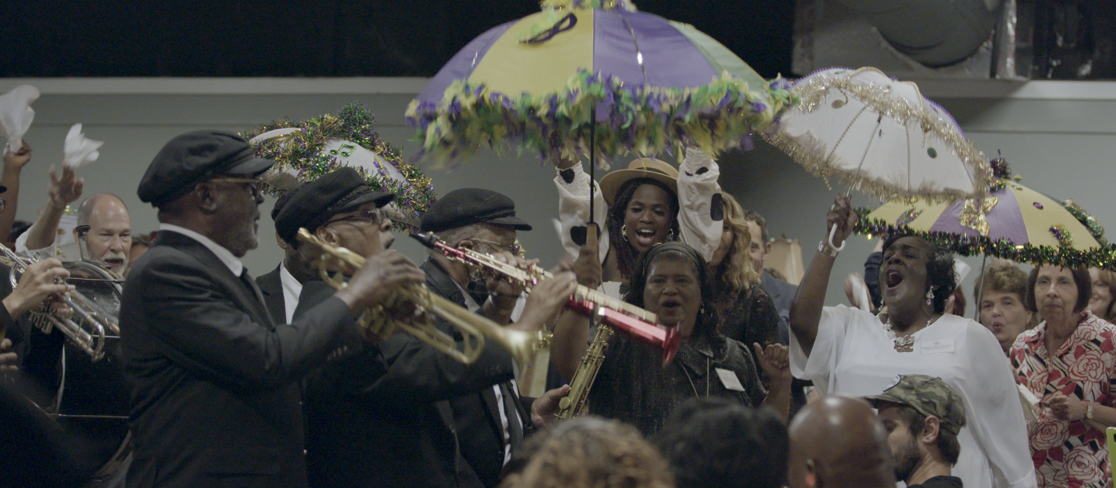 Musicians dressed in dark suits and caps walking through the street playing, with women holding colorful umbrellas cheering.