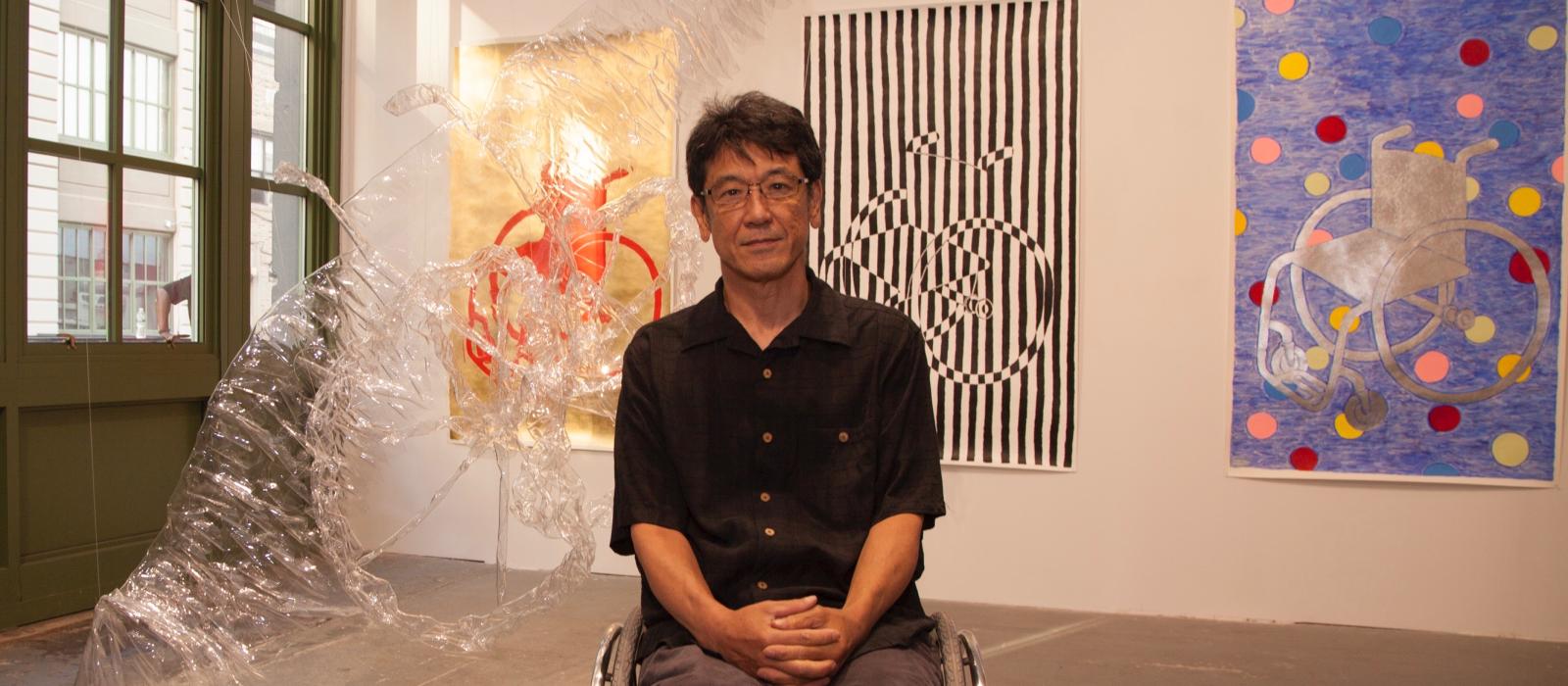 Man wearing glasses sitting in.wheelchair in front of a wall of paintings.