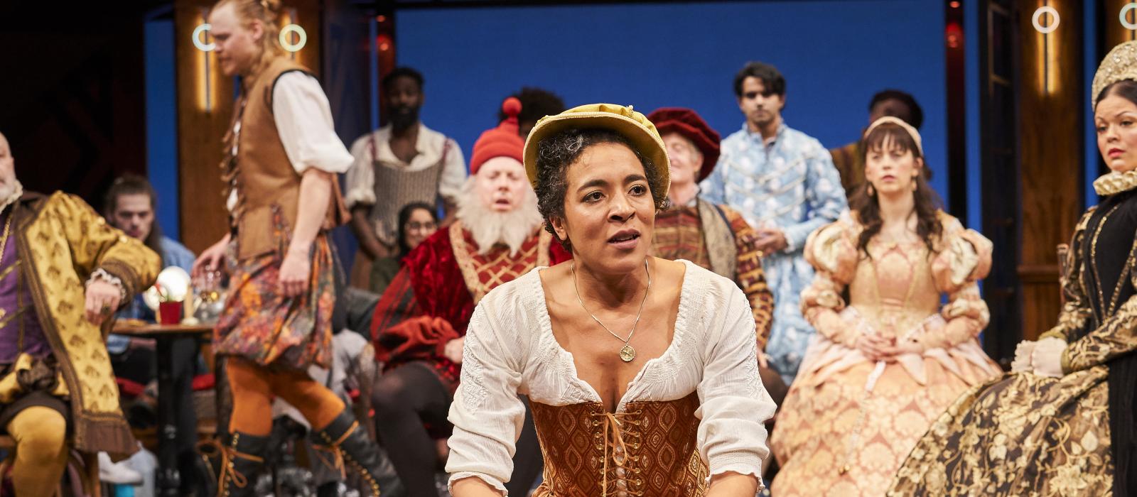 Woman in Shakespearean garb on stage in front of group of people in brightly colored clothes.