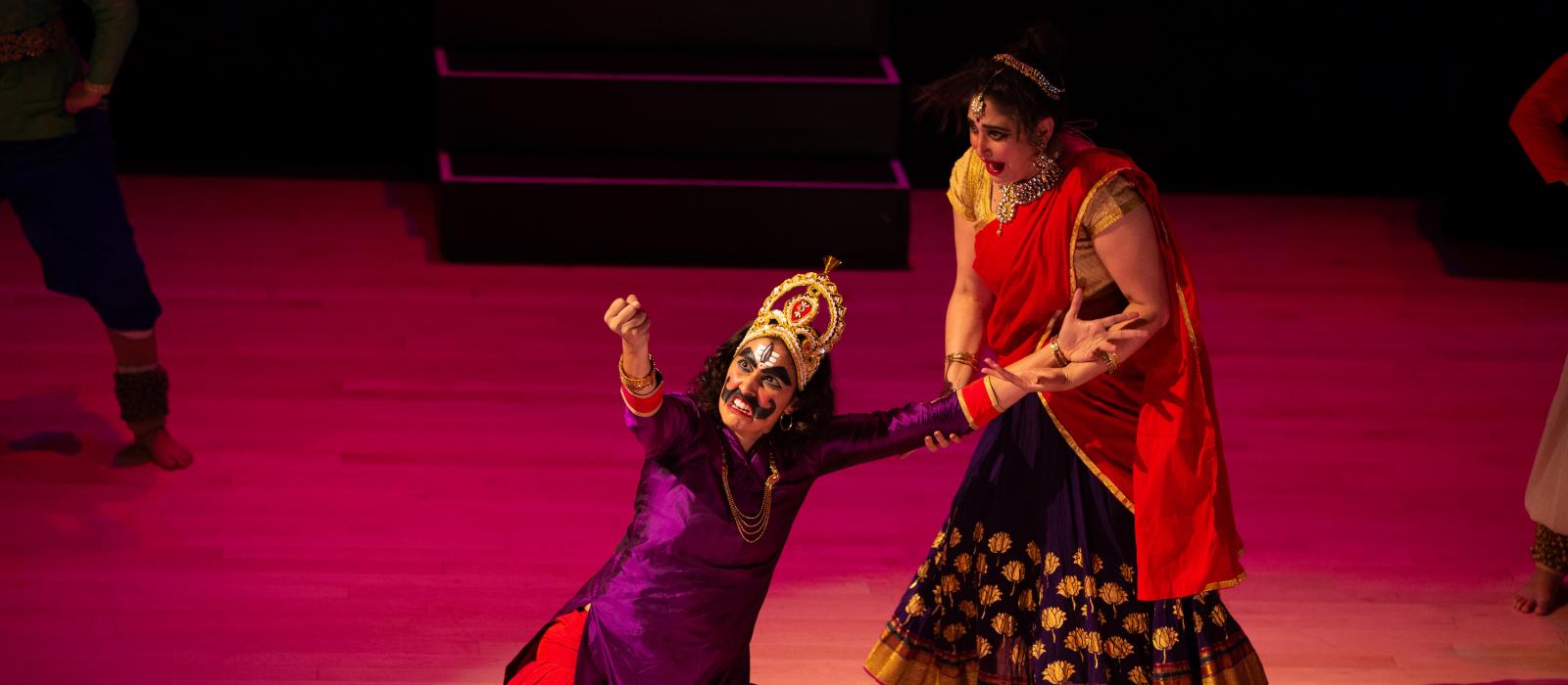 Stage production of two women in colorful clothing, one kneeling wearing fake moustache and shaking fist at sky and the other watching her.