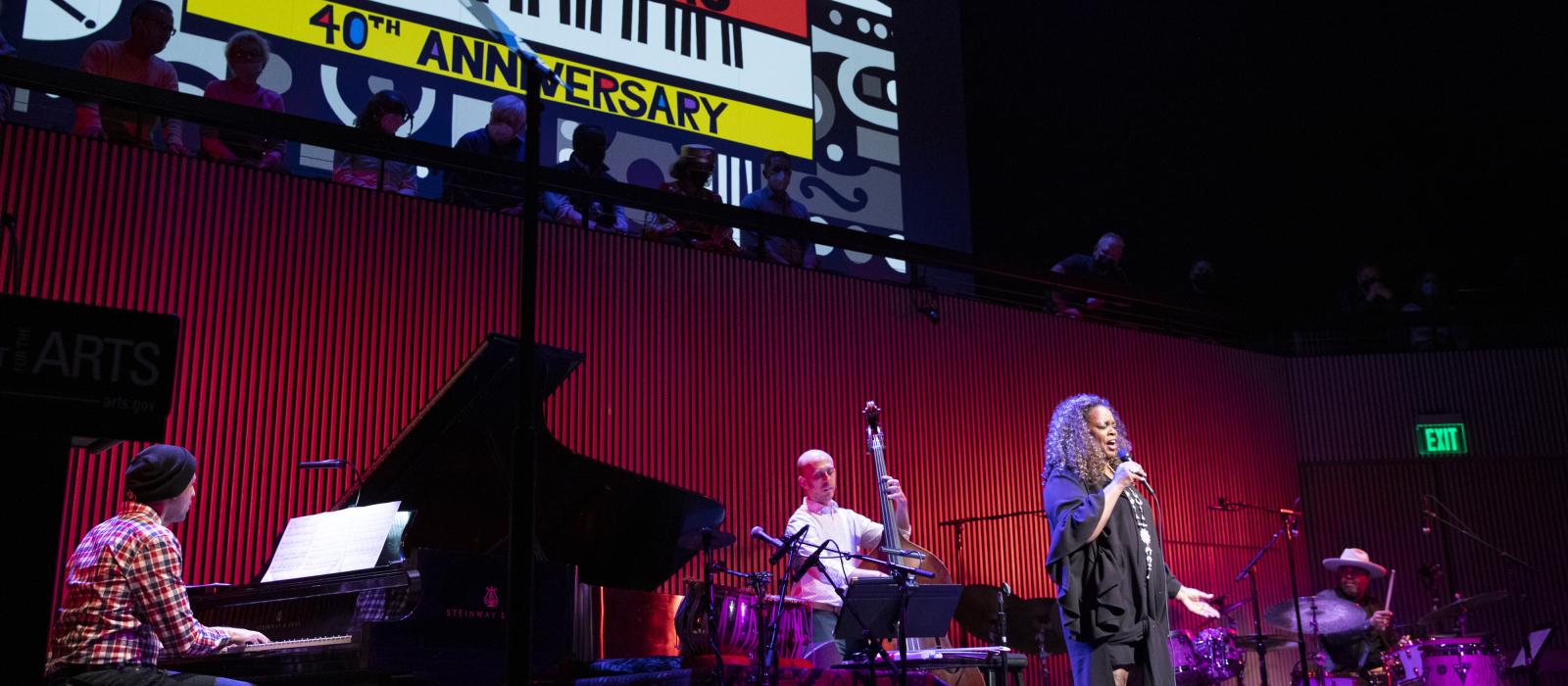 Black woman in black dress singing in front of a band on stage with a huge colorful banner behind them. 