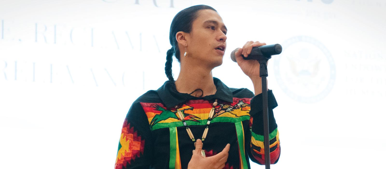 Native man with hair in braid, wearing colorful traditional clothes, at a microphone on stage.