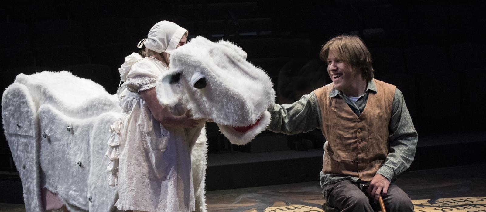 A man is seated on a stool, petting a large puppet of a cow. Another person dressed all in white hugs the cow.