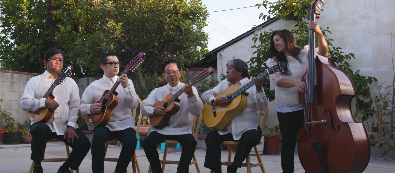 Group of musicians holding instruments seated outside all wearing white shirts.