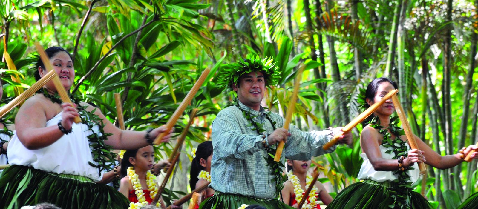Dancers wearing grass skirts and floral wreaths holding bamboo sticks perform a traditional Hawaiian dance.