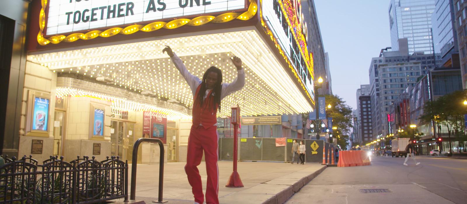 Black man wearing all red tap-dancing in front of a movie theater with the words "Together as One" on the marquee.