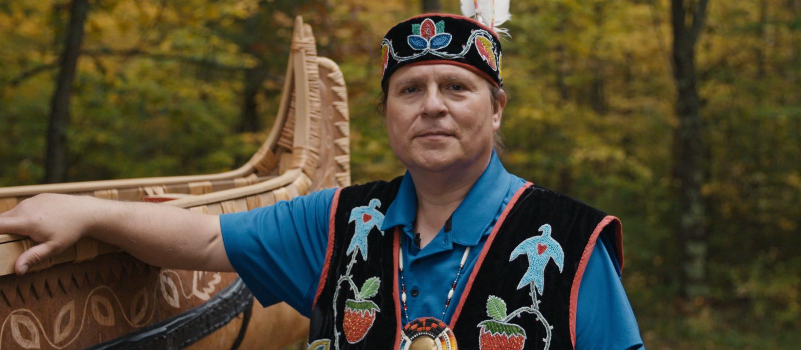 Native man with headband and feather wearing traditional clothes with an arm on a handbuilt canoe.
