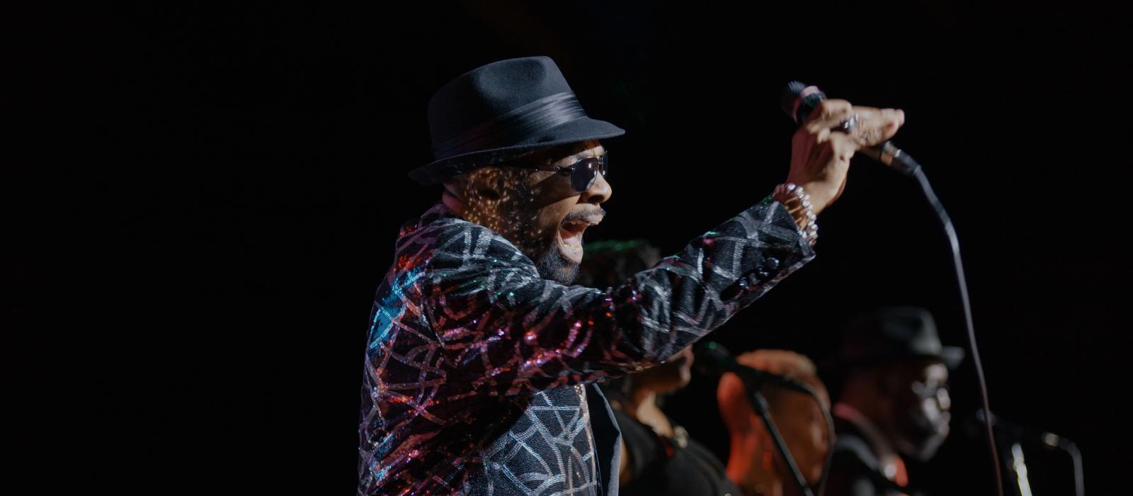 Black man in hat and shiny jacket singing into mic on stage.