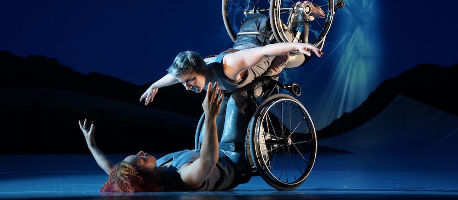 Laurel Lawson, a white woman with short cropped teal hair, is flying in the air with arms spread wide, wheels spinning, and supported by Alice Sheppard. Alice, a multiracial Black woman with coffee-colored hair, is lifting from the ground below. They are making eye contact and smiling. A burst of white light appears in a dark blue sky.