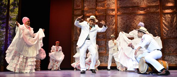 Man in white with white hat dancing on stage surrounded by musicians and other dancers, also dressed in white. 