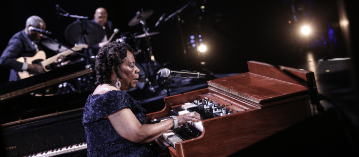 On a darkened stage, a woman plays an organ. A guitarist and drummer play in the background.
