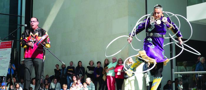 Woman in purple outfit dancing with hoops on stage while man to her right bangs and drum and sings.