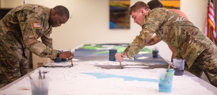 Two men in military uniforms bend over a long table from either side. They each are holding paintbrushes and are painting on a large sheet of paper that spans the length of the table between them.
