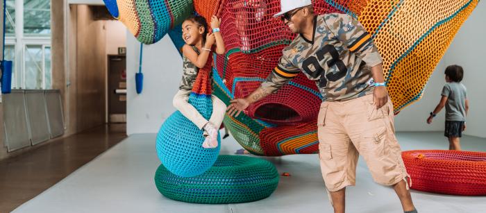 A man wearing a camouflage shirt with the number 93 and beige cargo shorts pushes a child sitting on a swinging blue netted ball, part of a colorful, large-scale netted play structure.