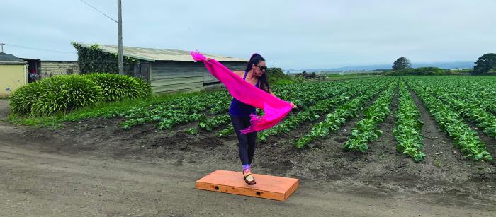 Woman with long dark hair wearing sunglasses dancing on a box with long orange scarf near a field of produce.