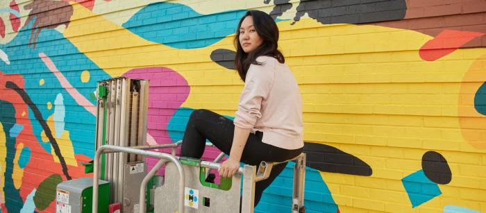 Asian women on scaffolding in front of mural she is painting. 