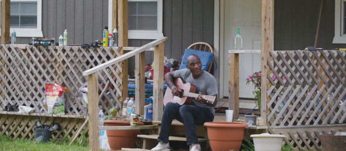 Black man wearing tee-shirt and jeans playing guitar on a porch of house. 