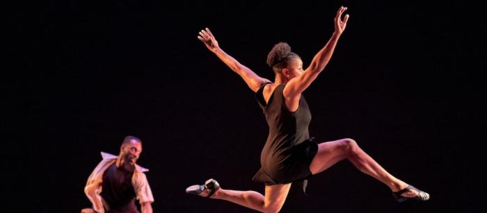 While a male-appearing dancer executes a move in the background, in the foreground a female-appearing dancer executes a leap.