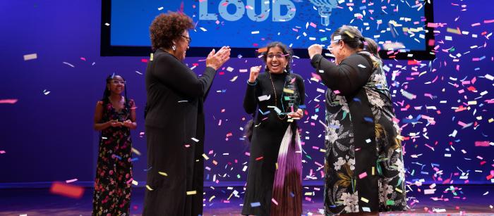 Two women of color clap on either side of a younger South Asian woman as confetti falls around them. 