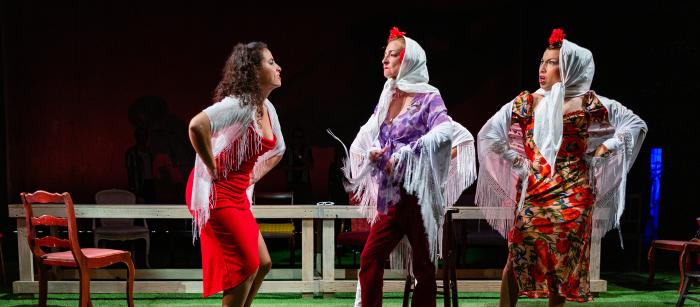 Woman in red dress and white shawl with hands on hips facing two older woman with red flower in their hair and white shawls over their head on stage.