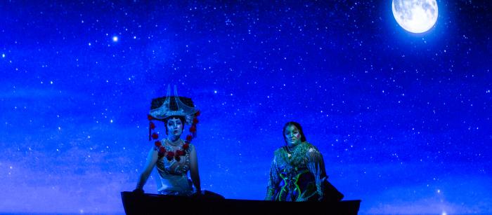 A stage production photo of two people ornately dressed in a boat with the moon behind them. 